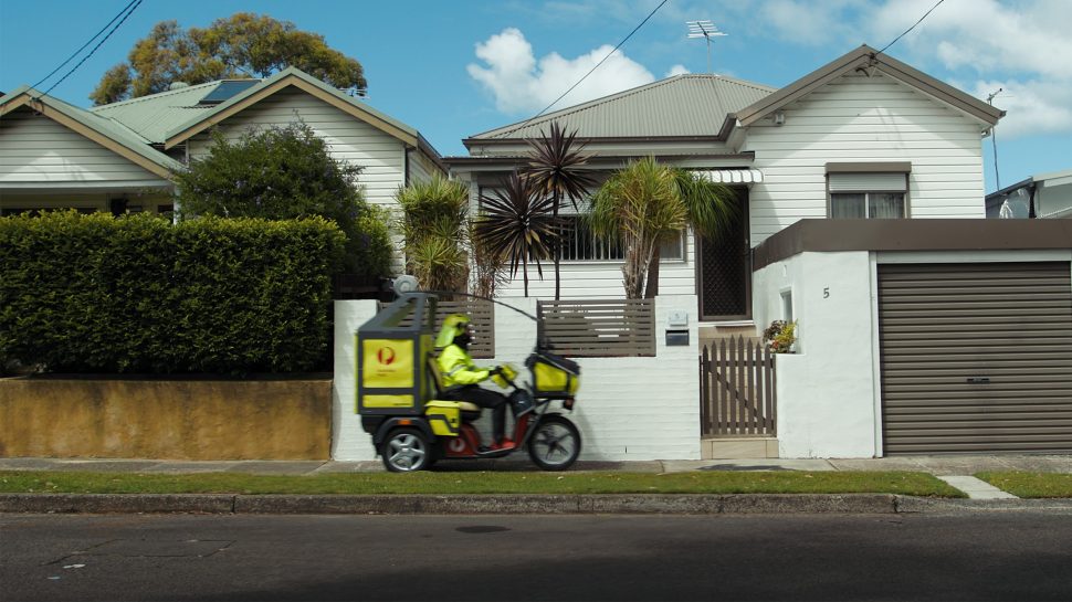 A Postie on an electric delivery vehicle drives along a suburban Australian street. 