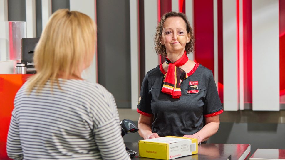 Female Australia Post postal staff with a physical disability is helping a customer send a parcel in a Post Office